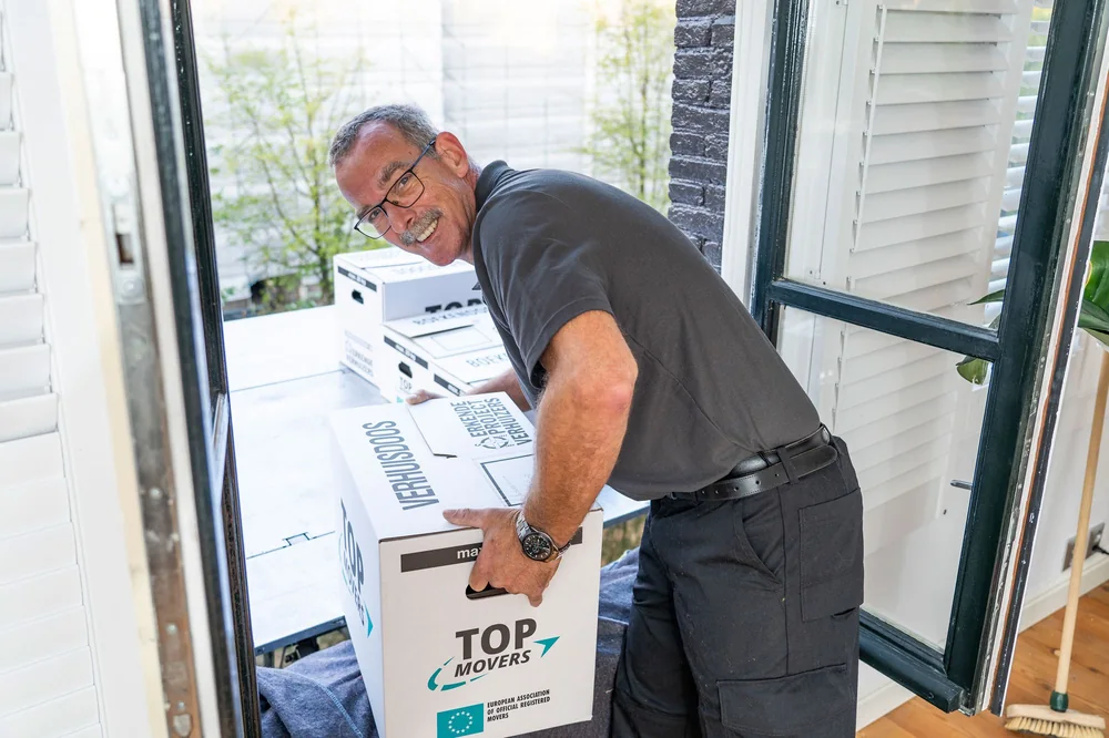 Verhuizer tilt een verhuisdoos van Erkende Projectverhuizers bij een open balkondeur