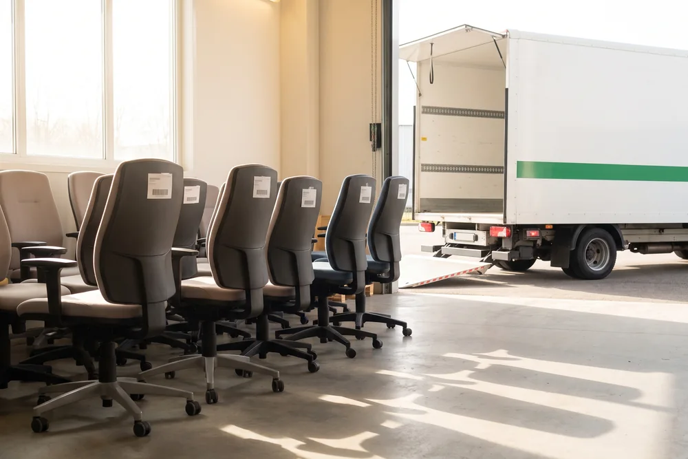 Rijen gebruikte bureaustoelen met labels in de opslaghal, met een vrachtwagen voor de open deur