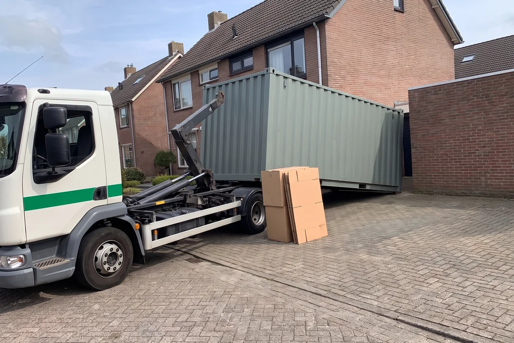 Witte containerwagen zet met de haakarm een opslagcontainer af op de oprit van een woning