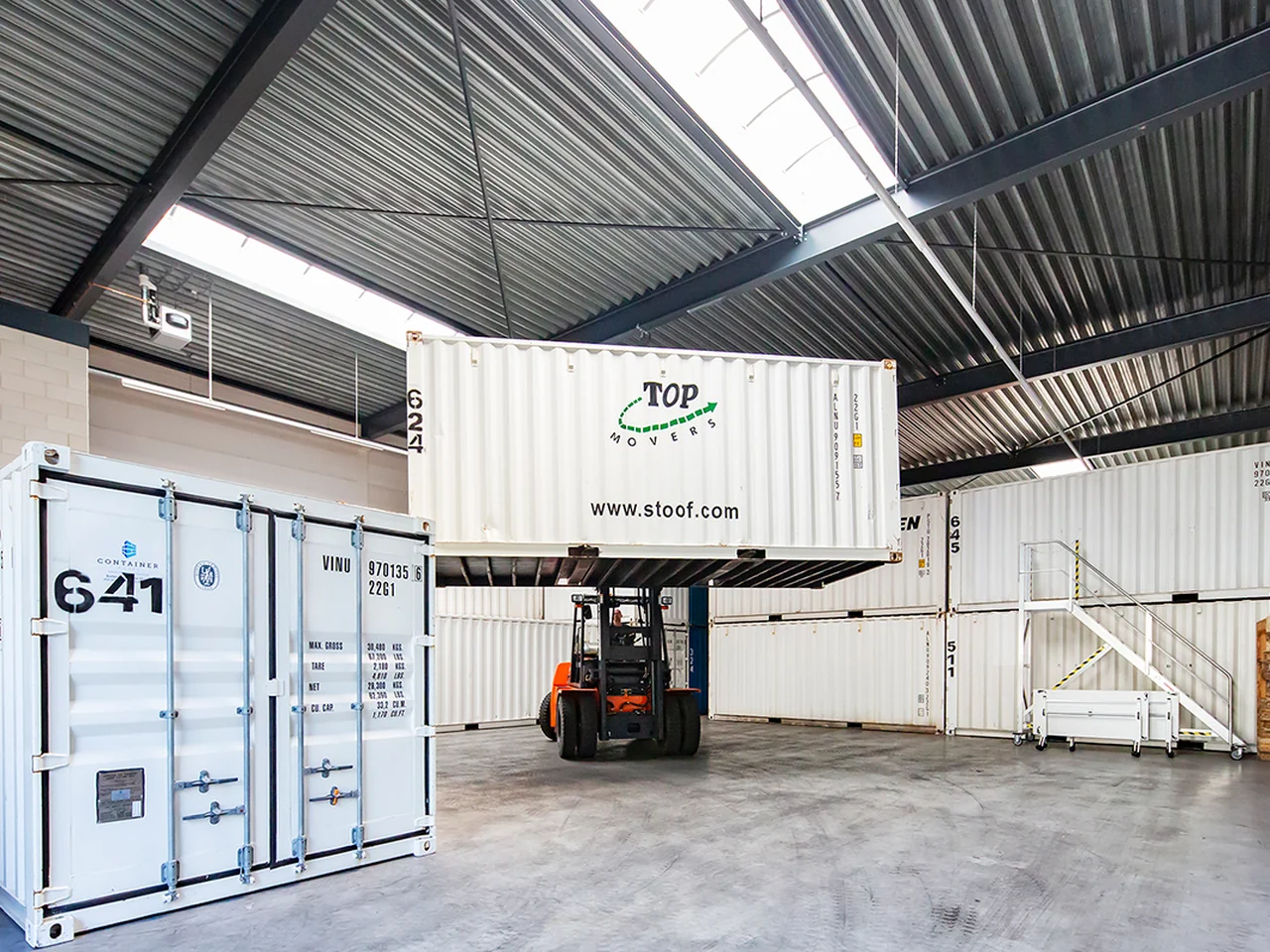 Heftruck tilt een container van Top Movers in een opslaghal met gestapelde containers