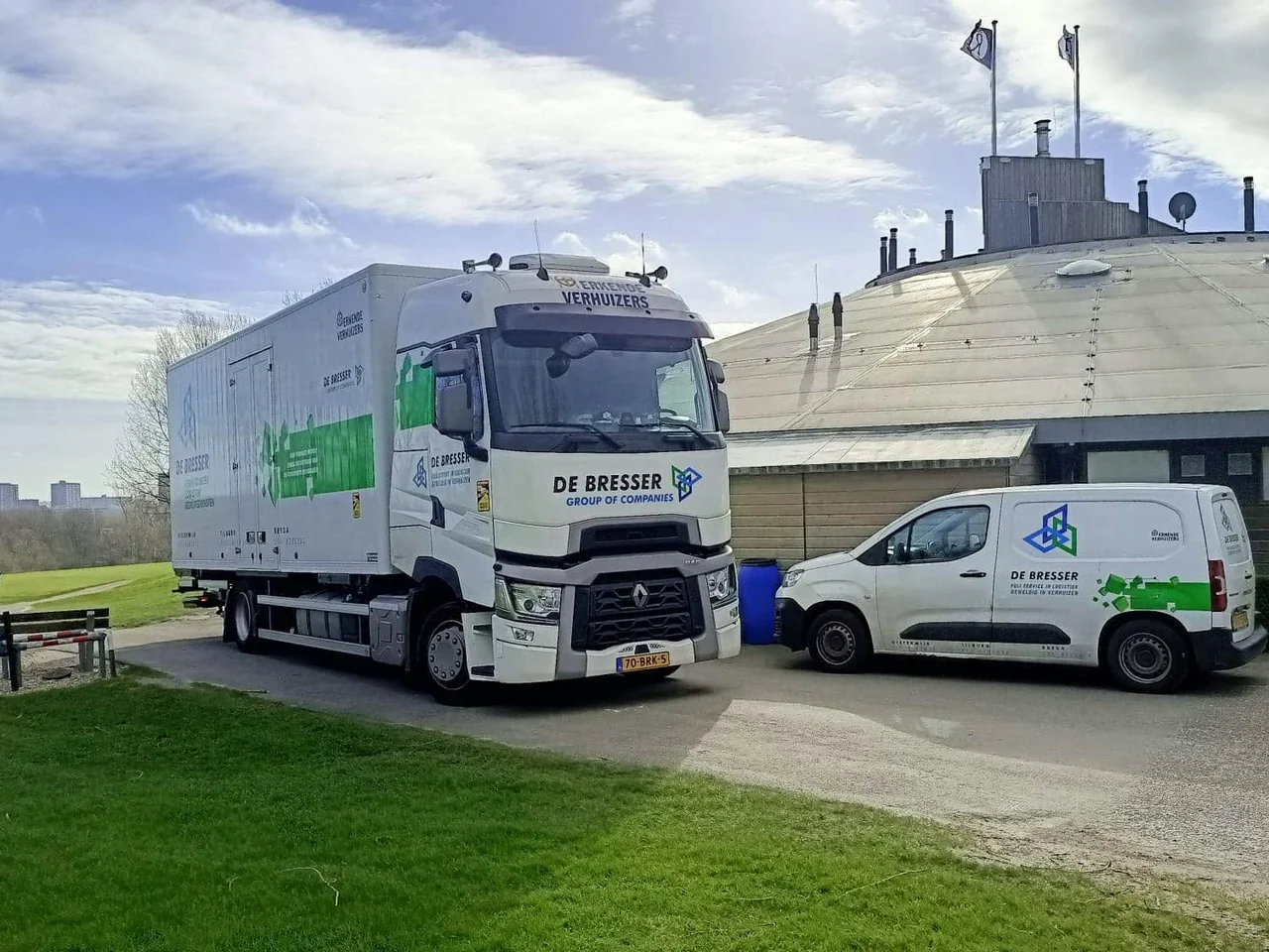 Vrachtwagen en bestelwagen van De Bresser geparkeerd bij een rond gebouw aan een grasveld