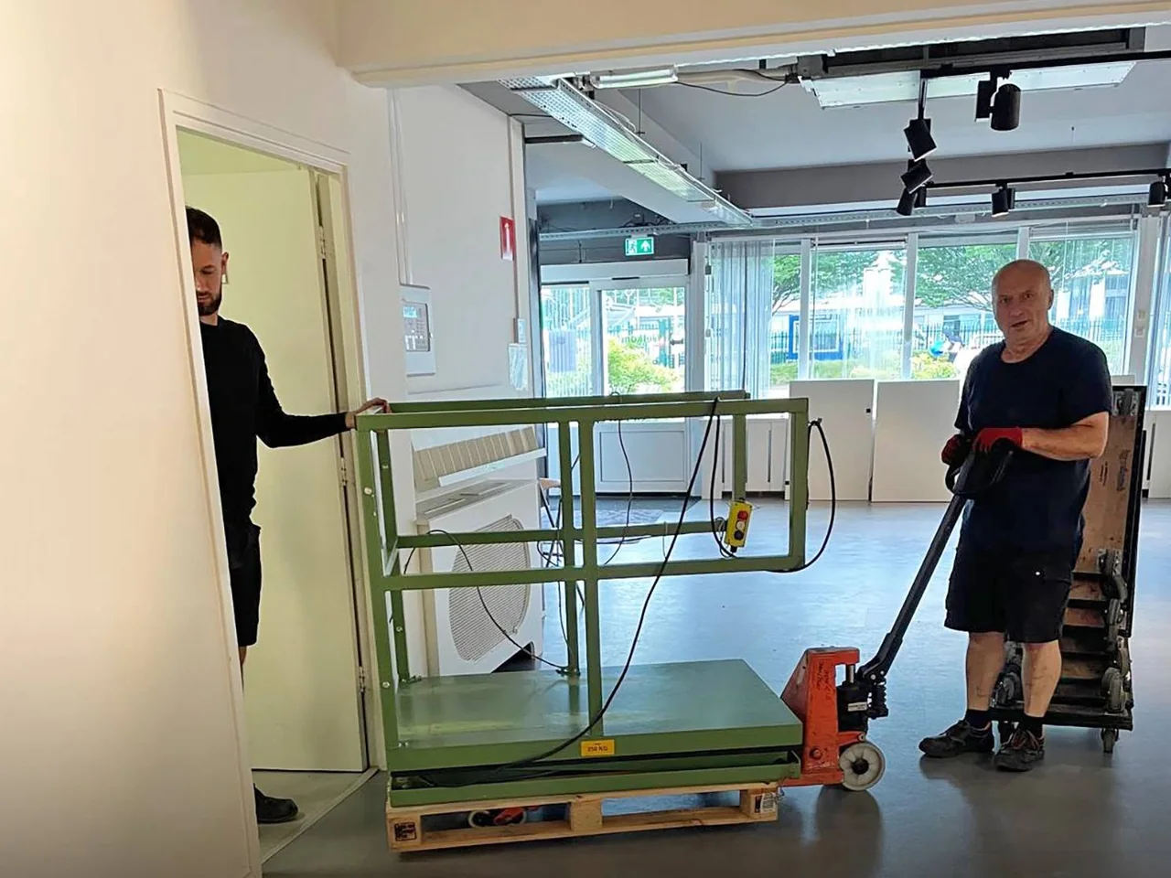 Twee medewerkers van De Bresser brengen een tafellift op een pallet met een palletwagen een lege bedrijfsruimte binnen