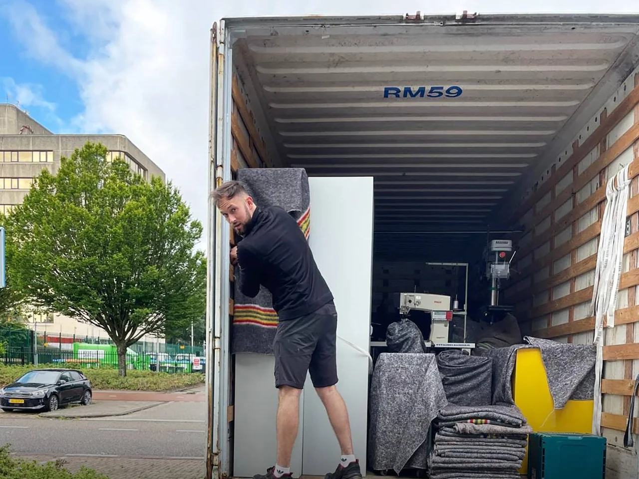 Medewerker van De Bresser zet een in een deken gewikkeld meubel in een deels geladen oplegger met machines en stapels verhuisdekens