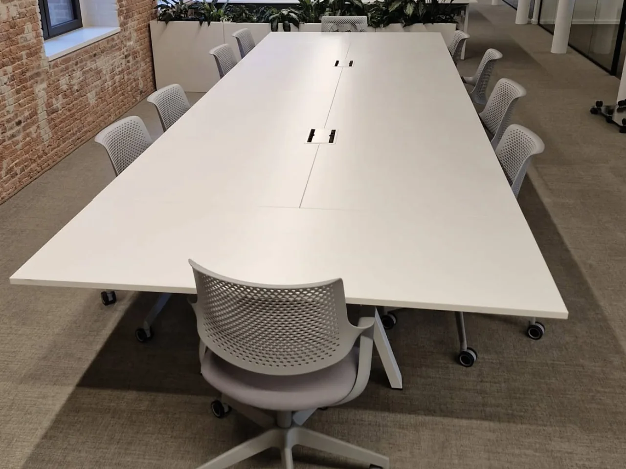 Nieuw ingericht kantoor met een lange vergadertafel, stoelen en planten langs een bakstenen wand