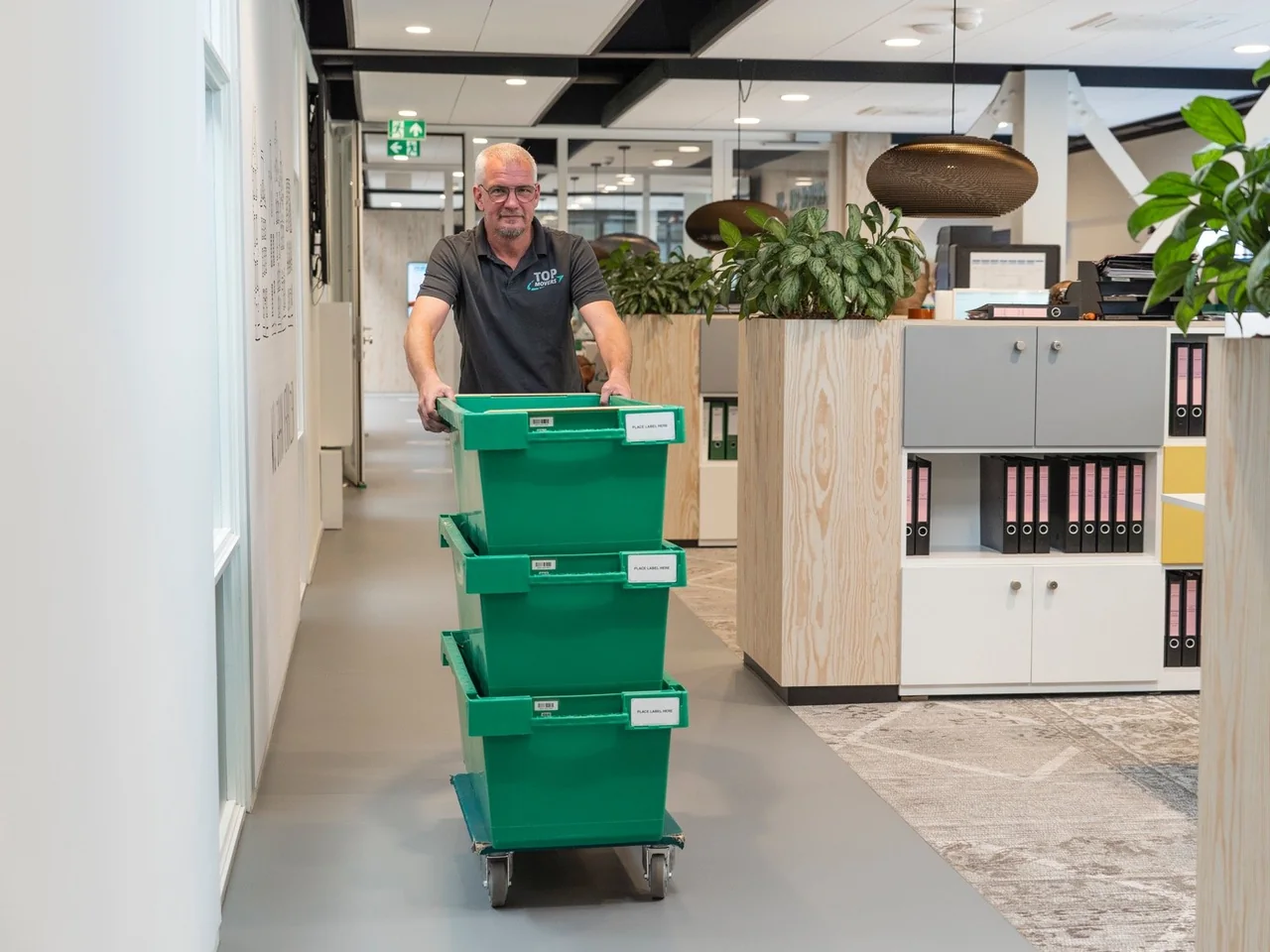 Verhuizer van De Bresser in Top Movers-polo rijdt groene verhuiskratten door een kantoorgang