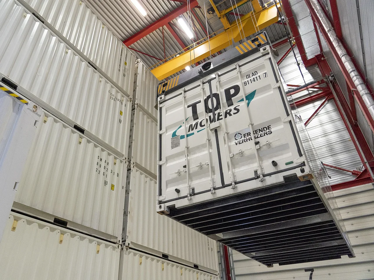 Container van Top Movers hangt aan een kraan tussen gestapelde opslagcontainers in een hal