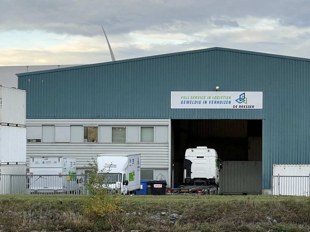 Blauwgroene loods van De Bresser in Tilburg met open laaddeur, een witte vrachtwagen binnen en containers ernaast