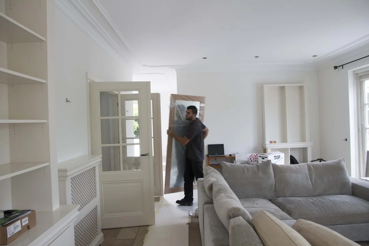 Verhuizer draagt een ingepakte spiegel door een lichte woonkamer met een grijze hoekbank