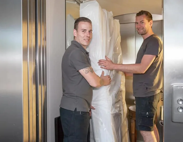 Twee verhuizers van De Bresser met een in folie verpakt meubelstuk in een lift